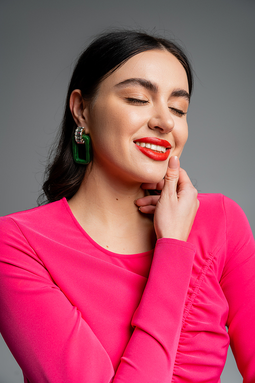 portrait of happy young woman with closed eyes, shiny brunette hair, trendy earrings, red lips and stylish magenta dress smiling while posing isolated on grey background