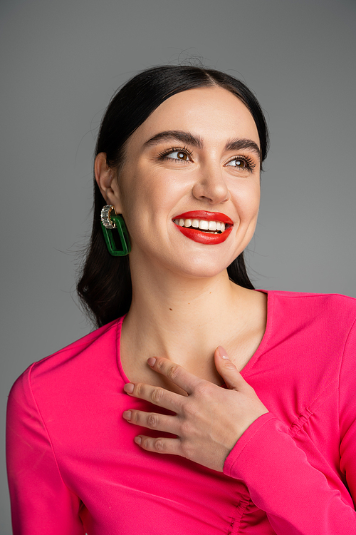 portrait of happy young woman with shiny brunette hair, trendy earrings, red lips and stylish magenta dress smiling while looking away and posing isolated on grey background