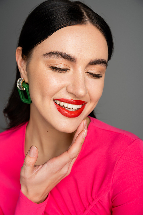 portrait of happy young woman with shiny brunette hair, trendy earrings and red lips smiling with closed eyes while posing isolated on grey background