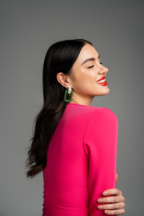 happy young woman with brunette hair, trendy earrings, red lips and stylish magenta dress smiling with closed eyes and posing on grey background