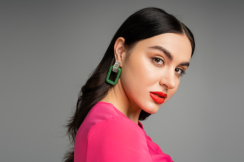 chic young woman with brunette hair, trendy earrings, red lips and stylish magenta dress looking at camera and posing on grey background