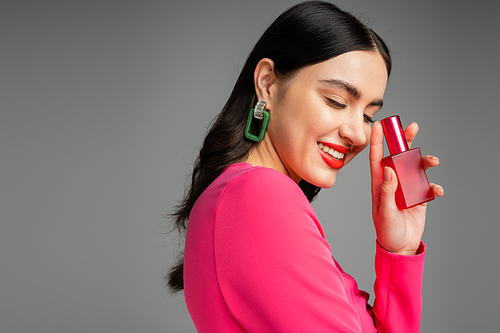 chic young woman with brunette hair, trendy earrings, red lips and stylish magenta dress holding bottle of luxurious perfume and smiling on grey background