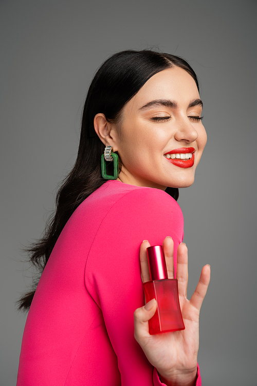 pleased woman with brunette hair, trendy earrings, red lips and stylish magenta dress holding bottle of luxurious perfume and smiling on grey background