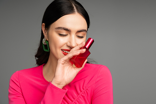 glamorous young woman with brunette hair, trendy earrings, red lips and stylish magenta dress holding bottle of luxurious perfume and smiling on grey background