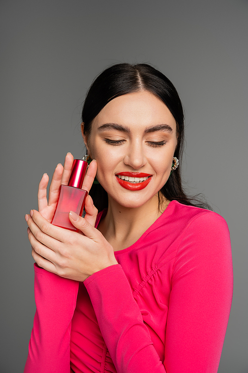 portrait of gorgeous young woman with brunette hair and red lips posing in magenta dress while holding bottle of luxurious perfume and smiling isolated on grey background