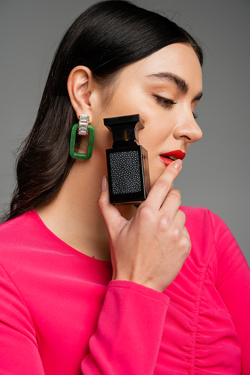 portrait of alluring young woman with brunette hair and red lips posing in trendy magenta dress while holding bottle of luxurious perfume isolated on grey background