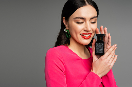 charming young woman with shiny brunette hair, red lips posing in magenta dress while holding bottle of perfume and smiling isolated on grey background