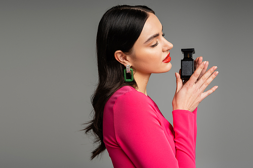 side view of alluring woman with shiny brunette hair, red lips and closed eyes posing in trendy magenta dress while holding bottle of luxurious perfume isolated on grey background