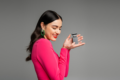 alluring young woman with shiny brunette hair, red lips posing in magenta dress while holding bottle of luxurious perfume and smiling isolated on grey background