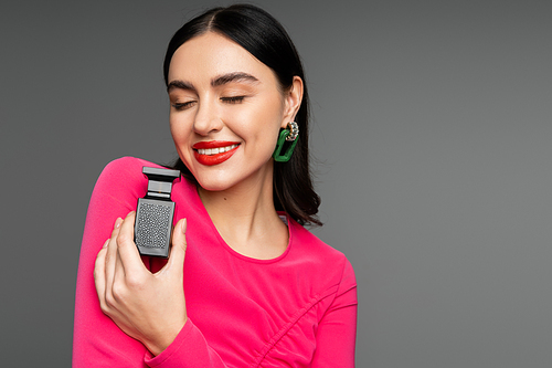 sophisticated young woman with shiny brunette hair, red lips, closed eyes and magenta dress holding bottle of perfume and smiling on grey background