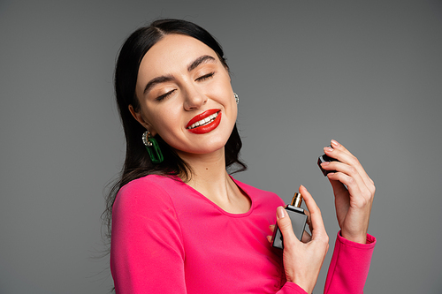 sophisticated young woman with brunette hair, red lips, closed eyes and magenta dress holding bottle and spraying luxurious perfume while smiling on grey background