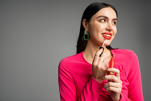 charming young woman with brunette hair and trendy earrings posing in magenta dress while applying red lip gloss on lips and smiling isolated on grey background