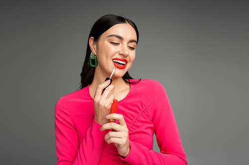 gorgeous woman with brunette hair and trendy earrings posing in magenta dress while applying red lip gloss before party isolated on grey background