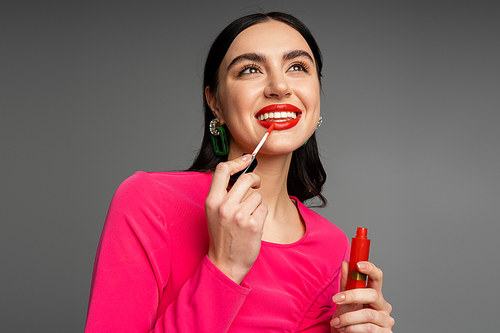 alluring young woman with shiny brunette hair and trendy earrings posing in magenta dress while applying red lip gloss before party isolated on grey background