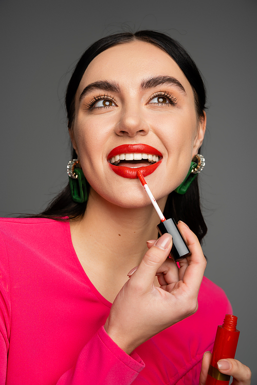 alluring woman with shiny brunette hair and trendy earrings applying red lip gloss and smiling while posing in magenta dress isolated on grey background