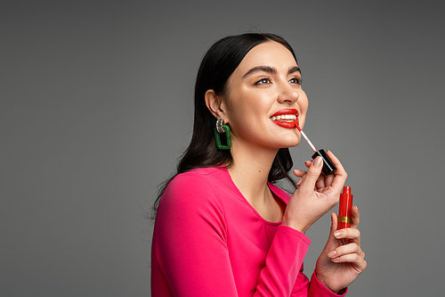 happy woman with brunette hair and trendy earrings applying red lip gloss and smiling while posing in magenta dress and preparing for party isolated on grey background