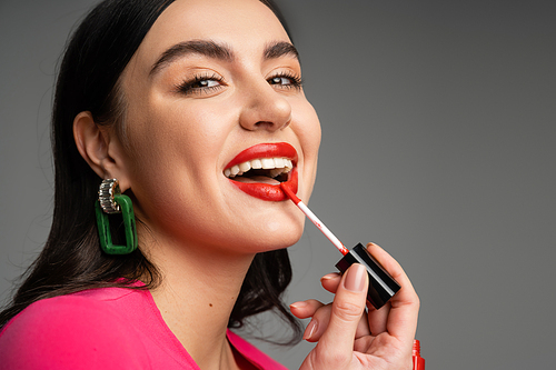 excited woman with shiny brunette hair and trendy earrings applying red lip gloss and smiling while posing in magenta dress isolated on grey background