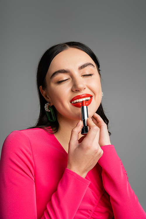 alluring woman with trendy earrings and shiny brunette hair applying red lipstick and smiling with closed eyes while and preparing for party isolated on grey background