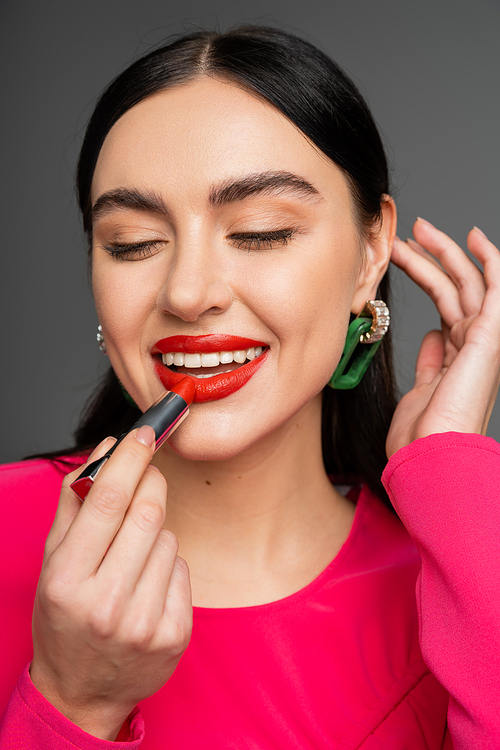 portrait of gorgeous woman with trendy earrings and brunette hair applying red lipstick and smiling while looking away and posing isolated on grey background