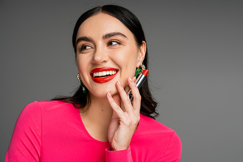 portrait of elegant woman with trendy earrings and shiny brunette hair holding red lipstick between fingers and smiling while posing on grey background