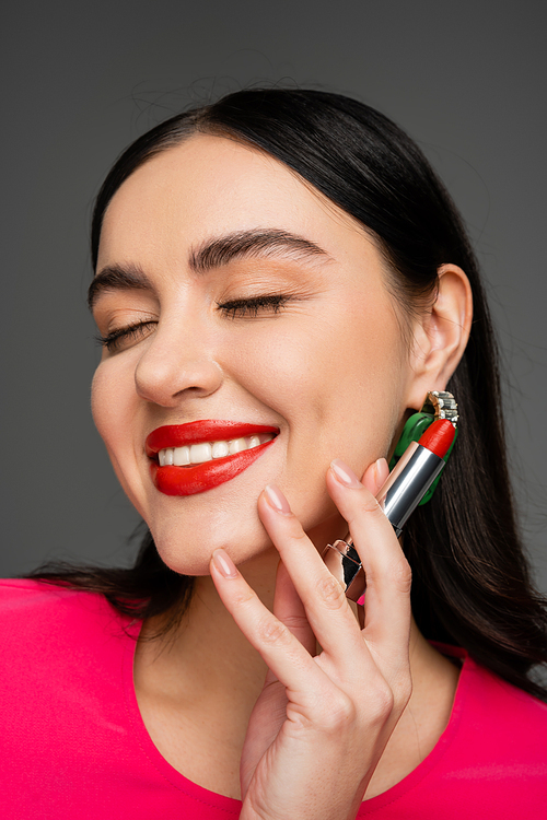 portrait of elegant woman with trendy earrings, flawless makeup and brunette hair holding red lipstick between fingers and smiling on grey background