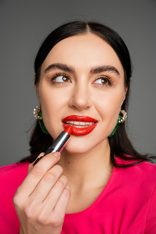 portrait of stunning young woman with trendy earrings and flawless makeup applying red lipstick while posing and looking away on grey background