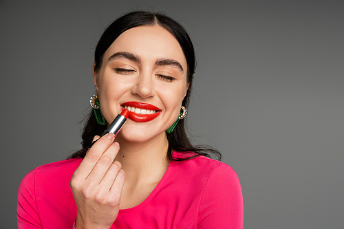 portrait of chic young woman with trendy earrings and flawless makeup applying red lipstick and smiling with closed eyes while posing on grey background