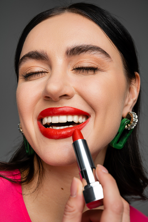 close up view of chic young woman with trendy earrings and shiny brunette hair and applying luxury red lipstick and smiling while posing on grey background