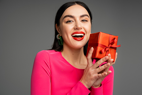 excited and chic young woman with trendy earrings and flawless makeup smiling while holding red and wrapped gift box on grey background