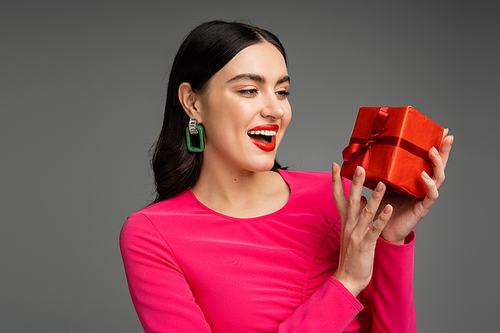 excited and chic young woman with trendy earrings and shiny brunette hair smiling while holding red and wrapped gift box on grey background