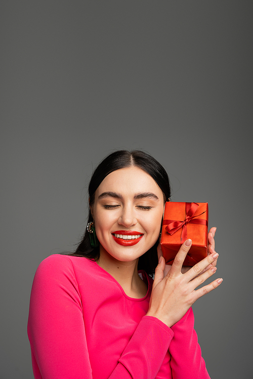 alluring and chic young woman with trendy earrings and shiny brunette hair smiling while posing with closed eyes and holding red wrapped gift box on grey background