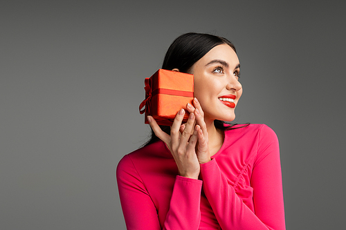 enchanting and chic young woman with trendy earrings and shiny brunette hair smiling while holding red and wrapped gift box on grey background with copy space