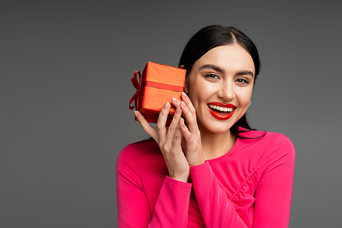 positive and chic young woman with trendy earrings and shiny brunette hair smiling while holding red and wrapped gift box on grey background