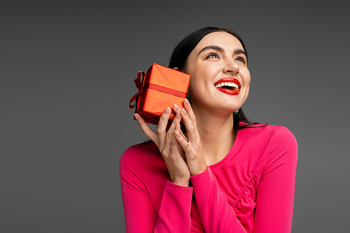 happy young woman with trendy earrings and shiny brunette hair smiling while holding red and wrapped gift box on holiday and looking up on grey background