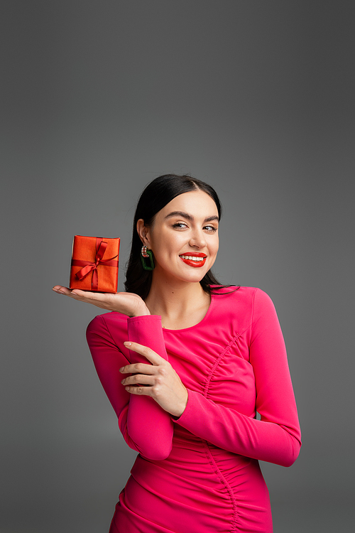 alluring and happy young woman with trendy earrings and shiny brunette hair smiling while holding red and wrapped present on grey background