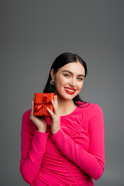 elegant and young woman with trendy earrings and brunette hair smiling while holding red and wrapped present for holiday on grey background