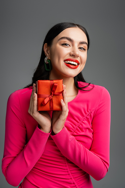 positive and young woman with trendy earrings and brunette hair smiling while holding red and wrapped present for holiday on grey background