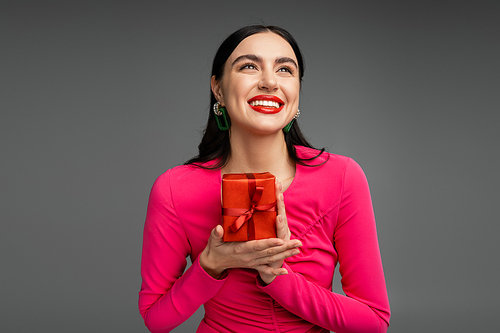 elegant and young woman with brunette hair and trendy earrings smiling while looking up and holding red and wrapped present for holiday on grey background