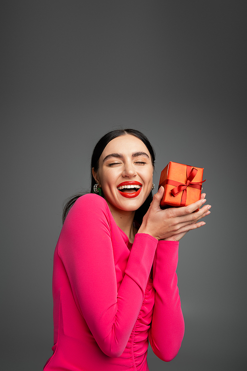 excited and young woman with trendy earrings and brunette hair smiling standing with opened mouth and holding red wrapped present for holiday on grey background