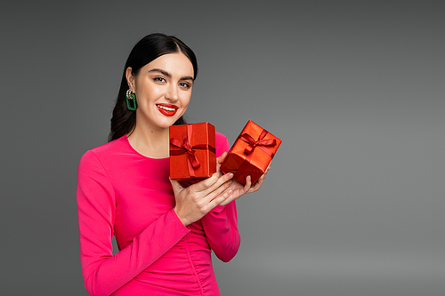 happy young woman with brunette hair smiling while standing in magenta party dress and holding wrapped gift boxes for holiday on grey background