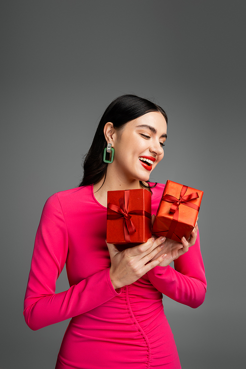 cheerful young woman with shiny brunette hair and trendy earrings smiling while standing in magenta party dress and holding wrapped gift boxes for holiday on grey background