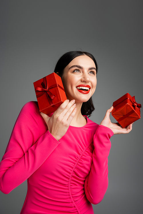 joyful young woman with brunette hair and trendy earrings smiling while standing in magenta party dress and holding wrapped gift boxes for holiday on grey background