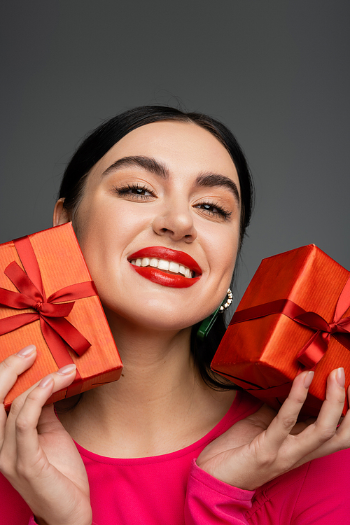 portrait of cheerful young woman with shiny brunette hair and trendy earrings smiling while holding wrapped gift boxes for holiday and looking at camera on grey background