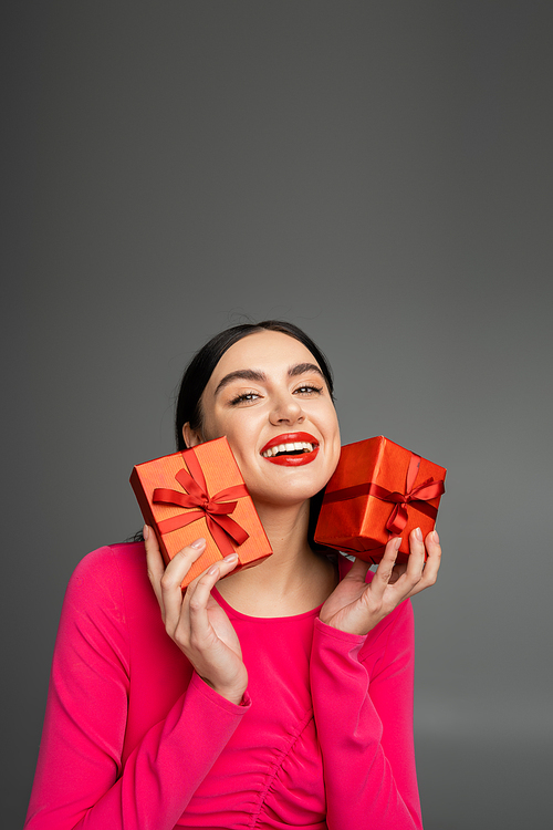 alluring young woman with shiny brunette hair smiling while standing in magenta party dress and holding wrapped gift boxes for holiday on grey background