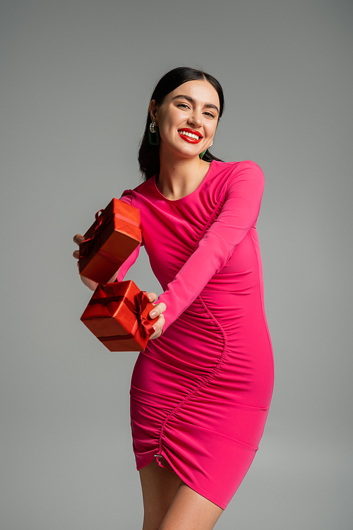 joyful and charming woman with brunette hair and trendy earrings smiling while standing in magenta party dress and holding wrapped gift boxes for holiday on grey background