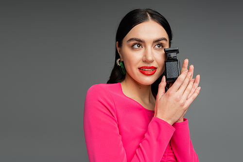 elegant and charming woman with brunette hair and trendy earrings standing in magenta party dress and holding bottle of perfume isolated on grey background