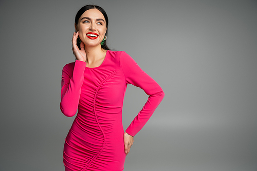 elegant and alluring woman with brunette hair and trendy earrings standing in magenta  party dress while posing with hand on hip and smiling isolated on grey background