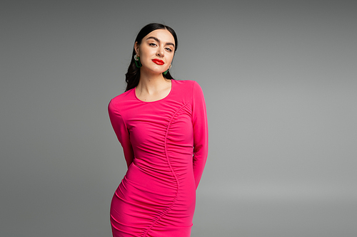 elegant and alluring woman with brunette hair and trendy earrings standing in magenta party dress and looking at camera isolated on grey background