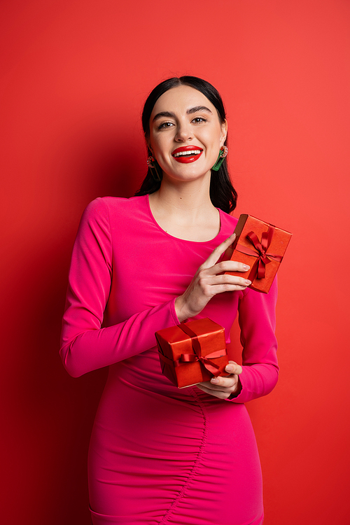 joyful and charming woman with brunette hair and trendy earrings smiling while standing in magenta party dress and holding wrapped gift boxes for holiday on red background