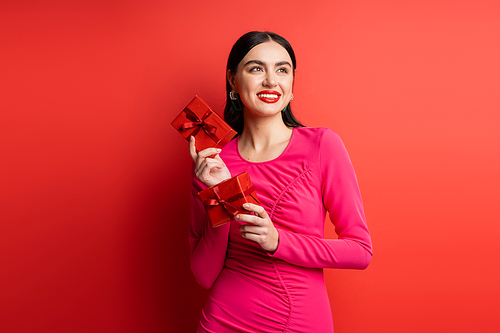 cheerful and elegant woman with brunette hair smiling while standing in magenta party dress and holding wrapped gift boxes for holiday on red background